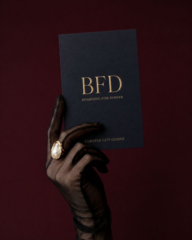 Hand in a sheer black glove holding a navy Boarding for Dinner guide against a burgundy backdrop