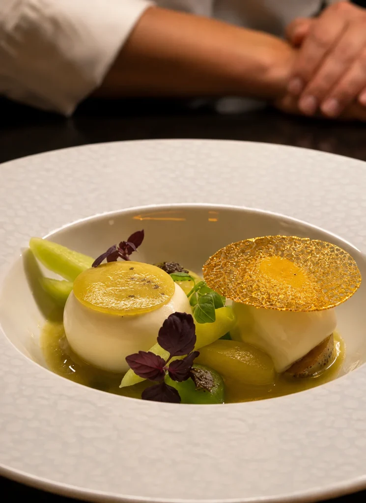Plated dish with golden-topped discs, green sauce and purple herbs in a white bowl