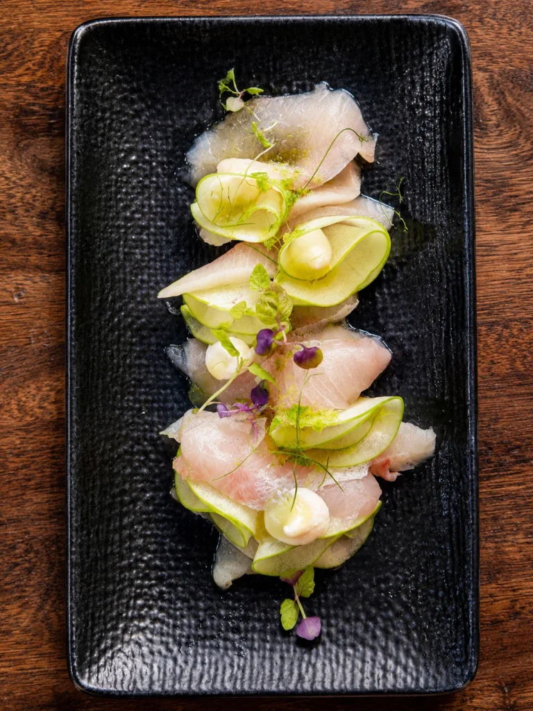 Sliced white fish crudo with lime and edible flowers on a black plate